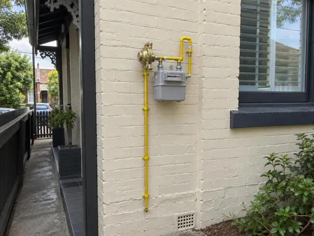 Exterior gas meter with yellow pipework on a cream brick house wall, beside a window in Leichhardt